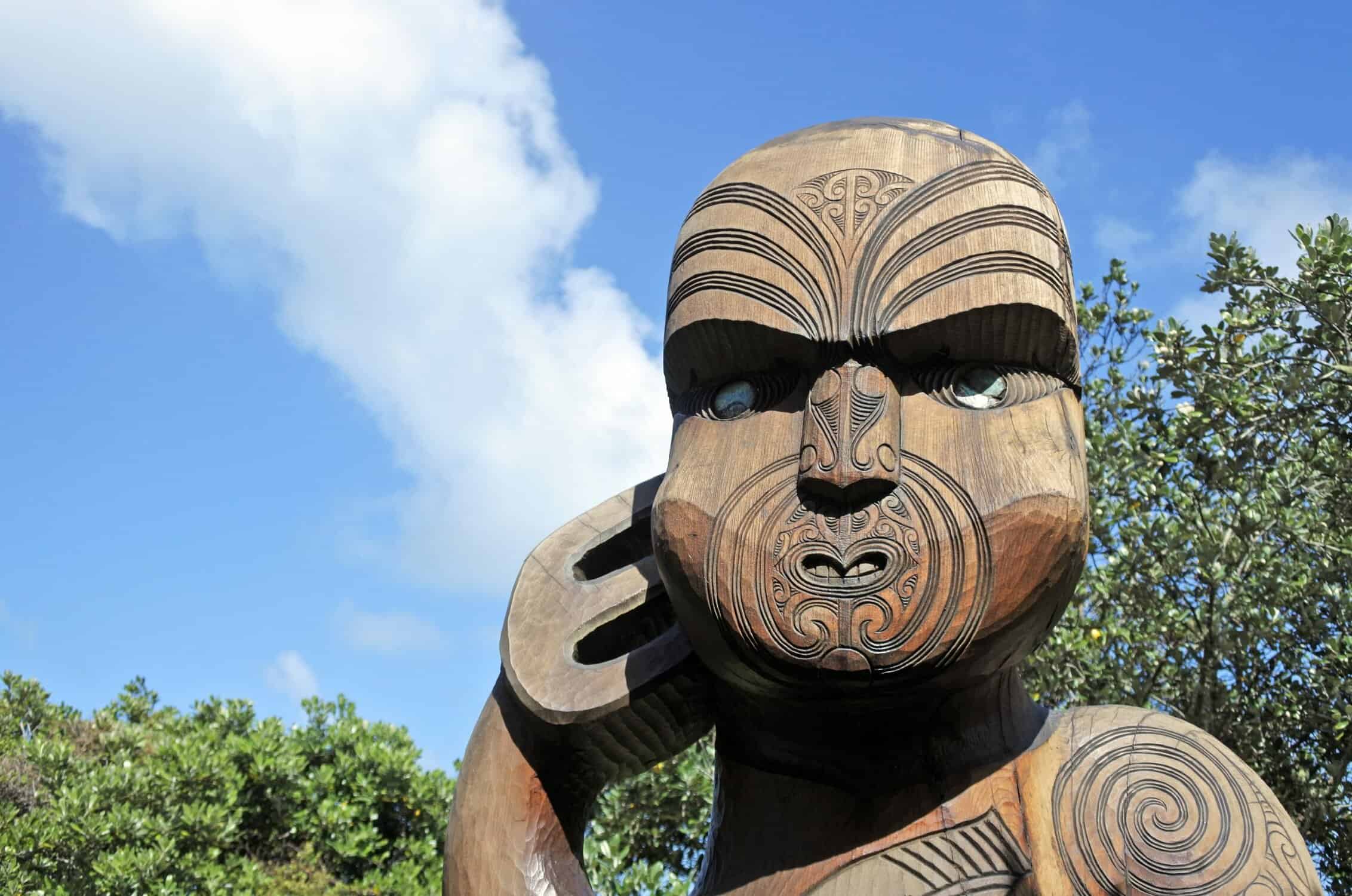 A carved wooden Māori figure with detailed moko, photographed against a blue sky